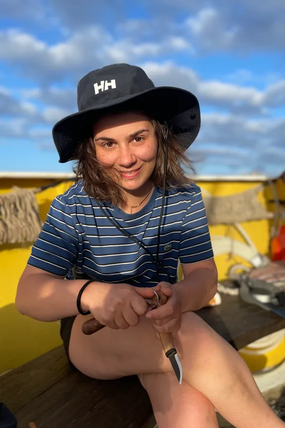 Alice Cherki zit aan dek in een gestreept shirt en zonnehoed, lachend, terwijl ze met een mes een houten lepel snijdt.