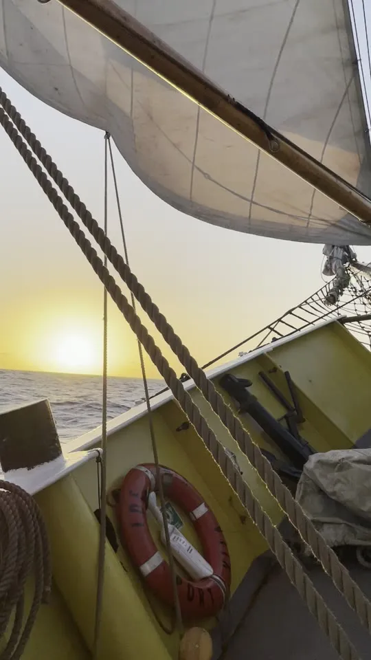 A wooden cask lashed on the sunlit deck beside coiled rope and yellow bulwarks, sails drawing overhead, the sun low on the horizon