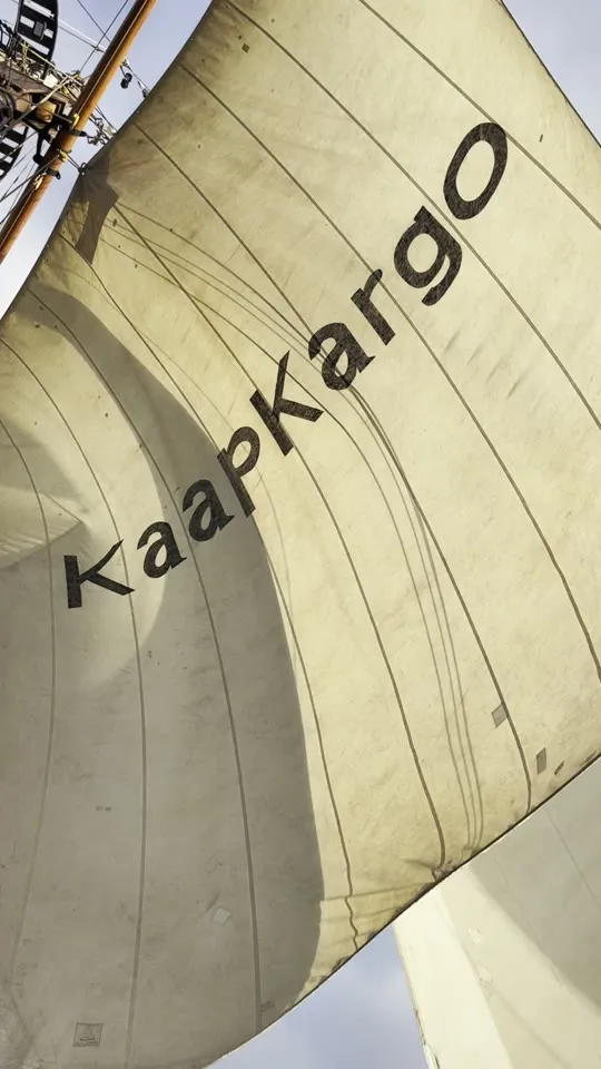 Looking up at Ide Min's sail filled with wind, KaapKargo lettered large across the canvas against a pale sky