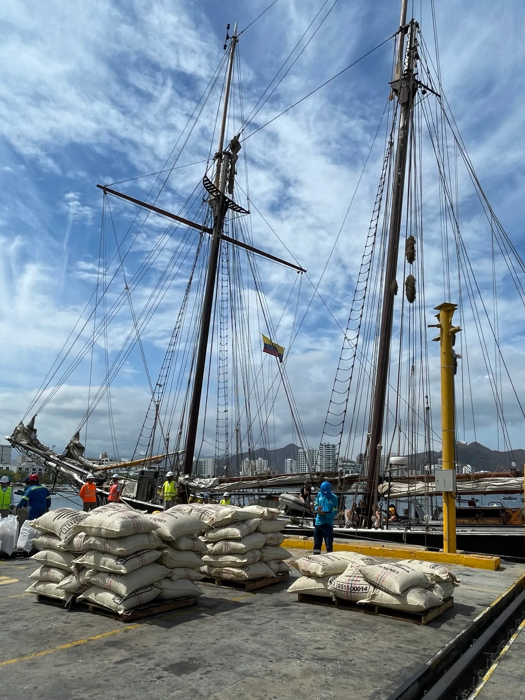 Coffee sacks stacked dockside with the ship's masts rising behind them and a Colombian flag visible