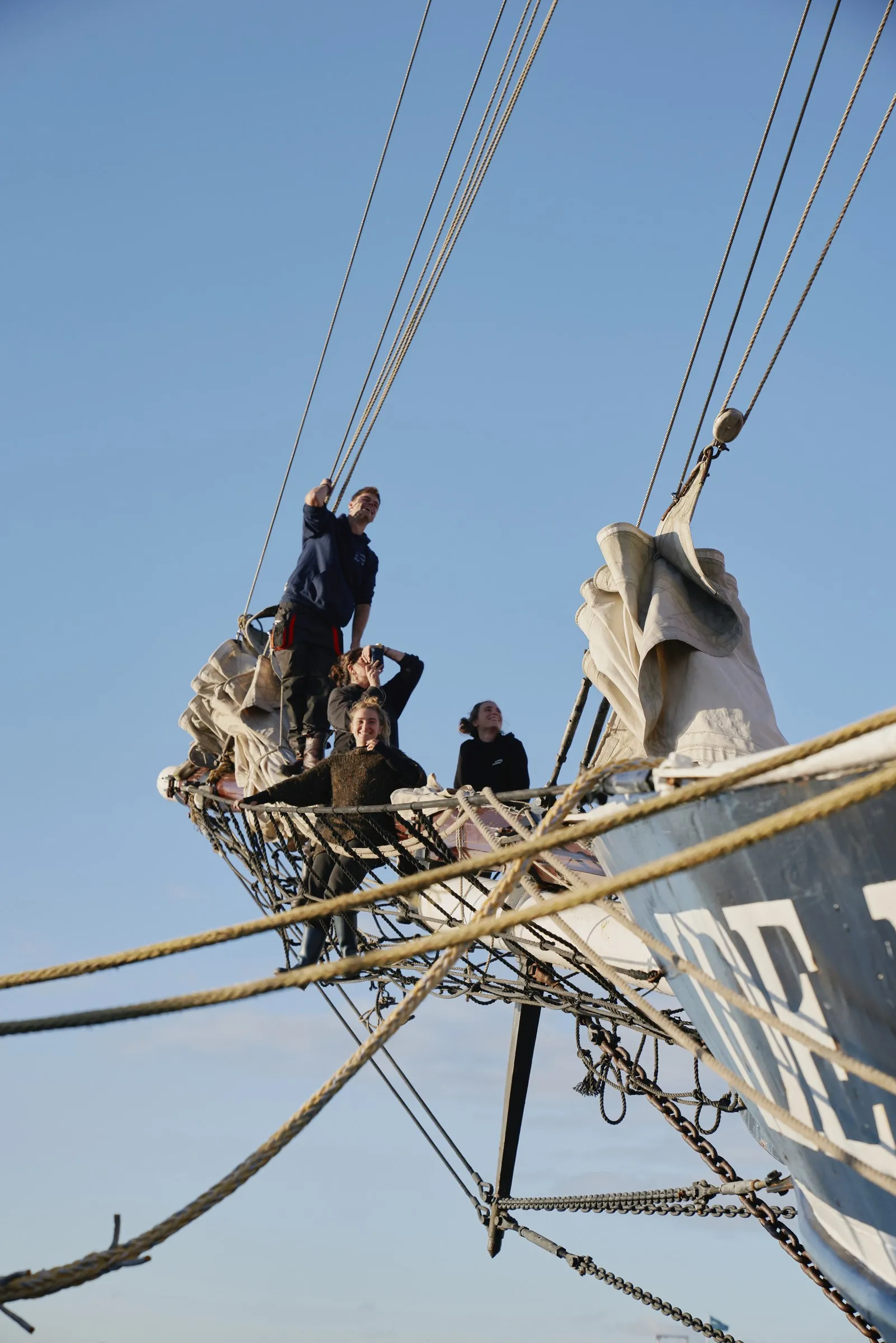 Vier bemanningsleden in het net van de boegspriet tegen een heldere lucht, één staand aan de stag, met de opgeborgen voorzeilen naast zich