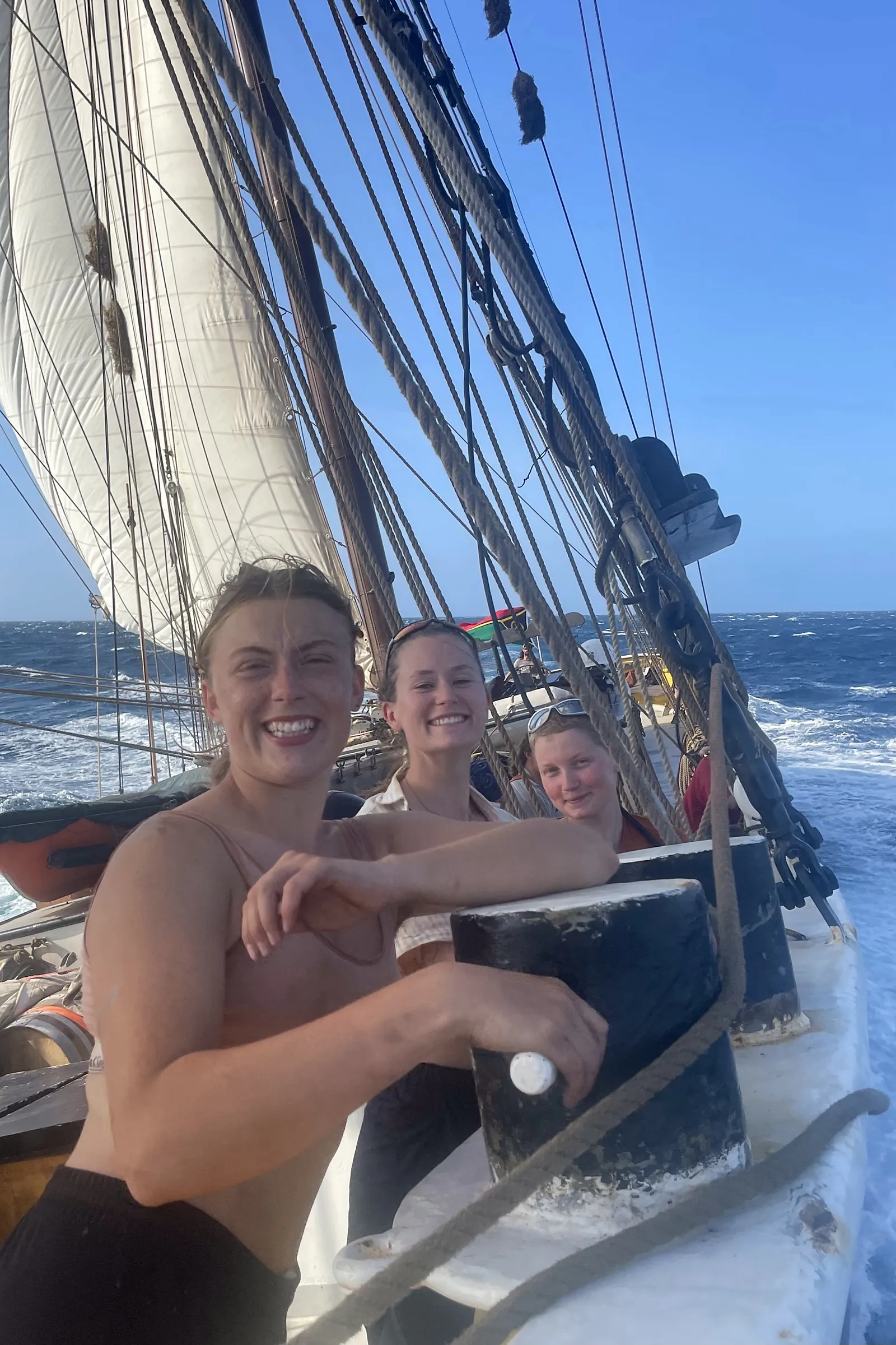 Drie bemanningsleden lachend bij de reling terwijl het schip onder vol zeil door open blauw water helt, met het kielzog ernaast