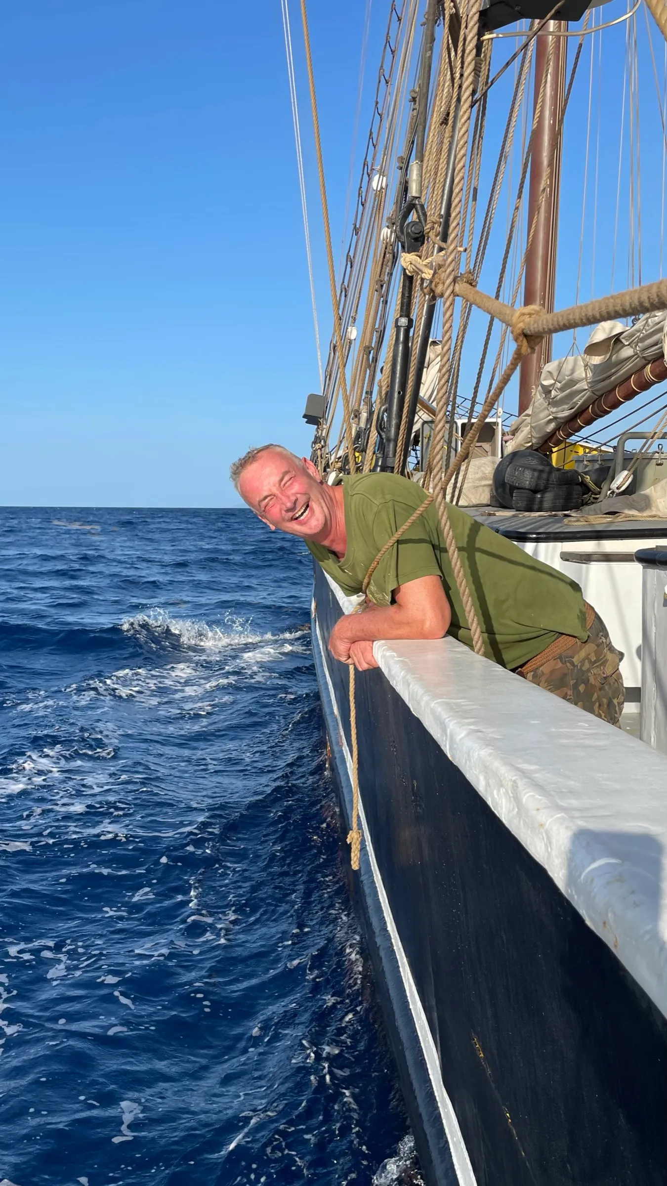 An older crew member leaning on the rail and laughing, deep blue open water and the ship's wake running past below, rigging and furled sail behind