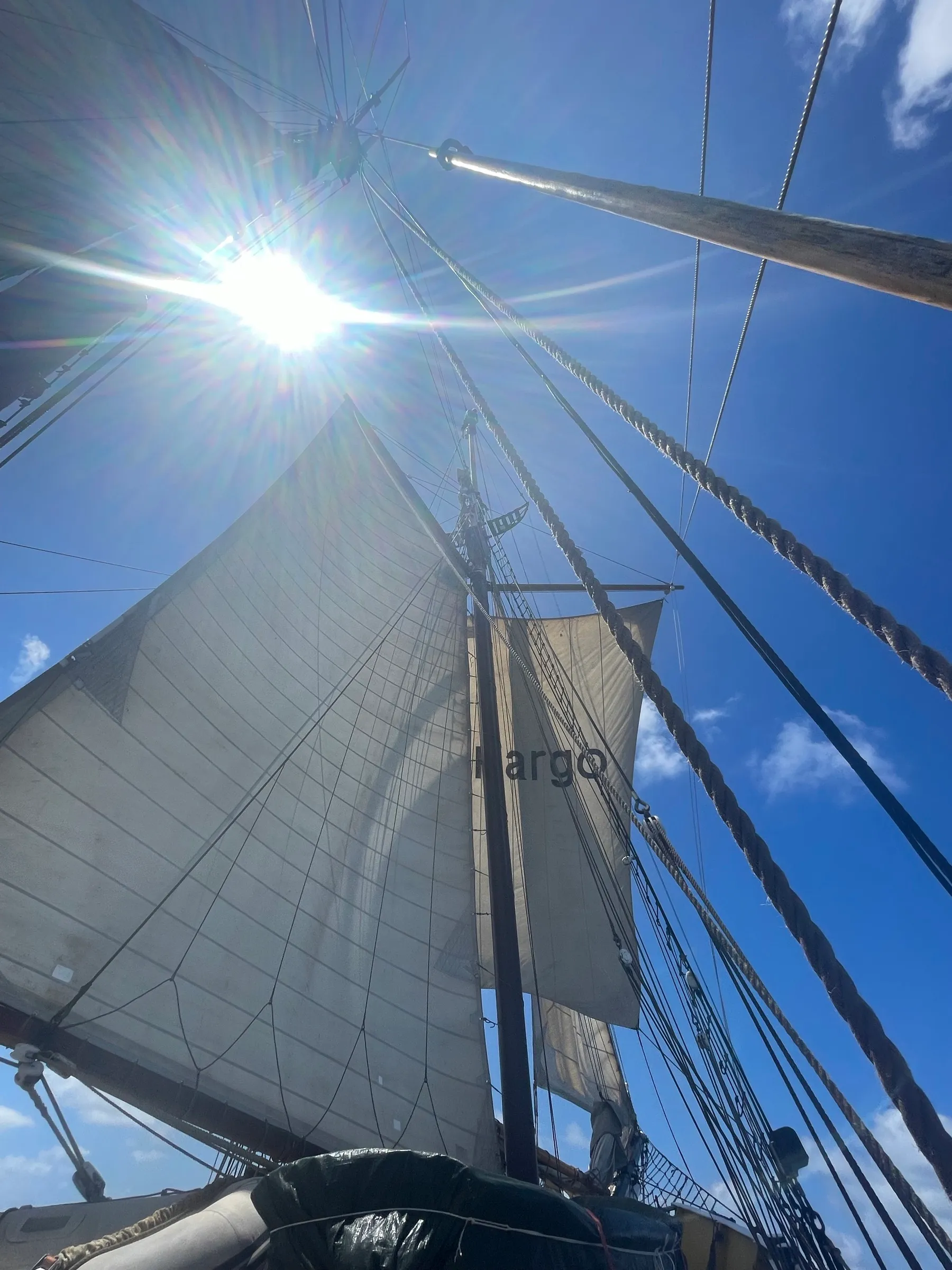 Vanaf het dek omhoog gekeken naar de volle zeilen van Ide Min tegen een blauwe lucht, met de zon door het tuig