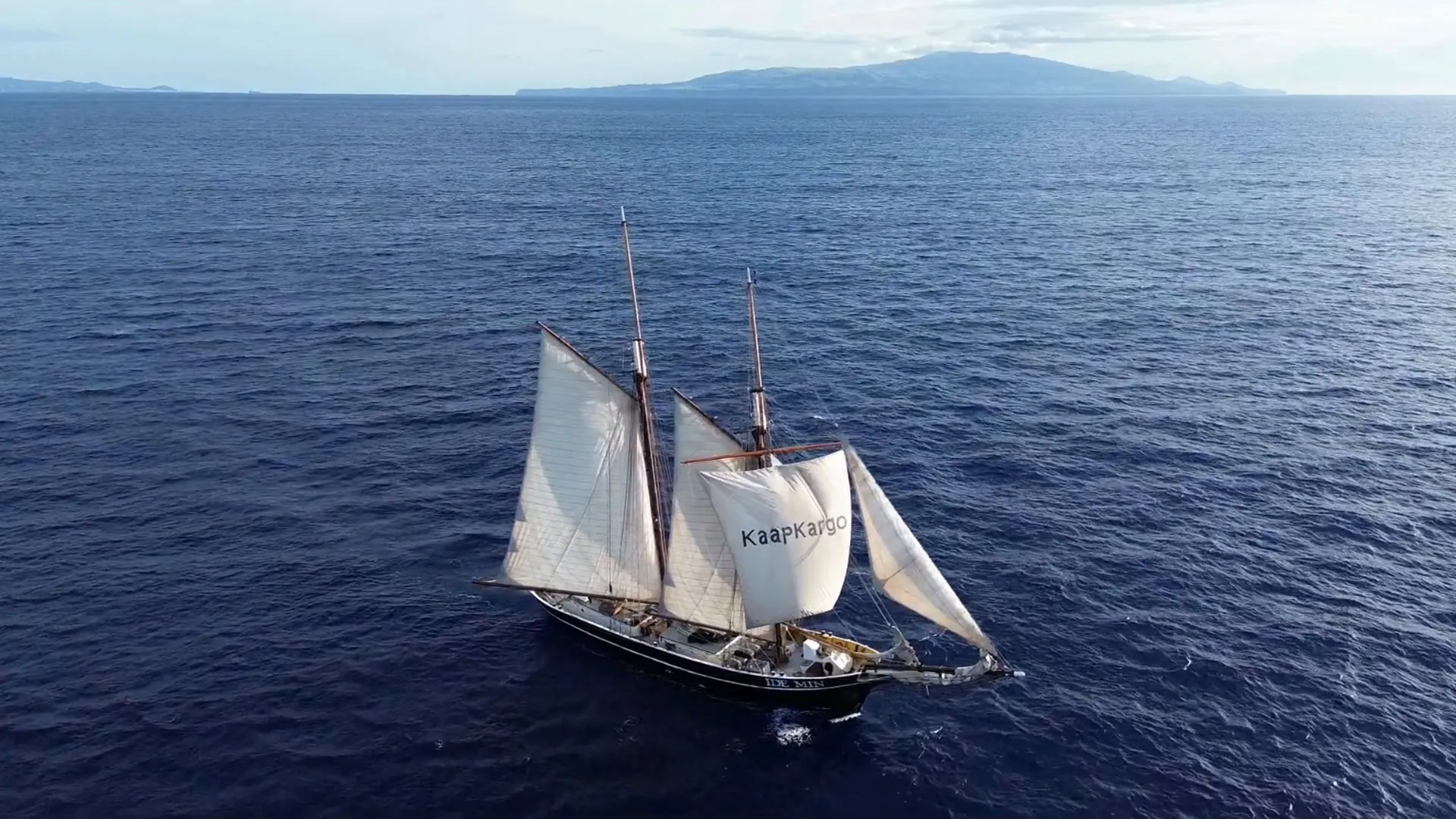 Ide Min under full sail on a deep blue sea seen from the air, Kaap Kargo lettered on her foresail, a mountainous island on the horizon