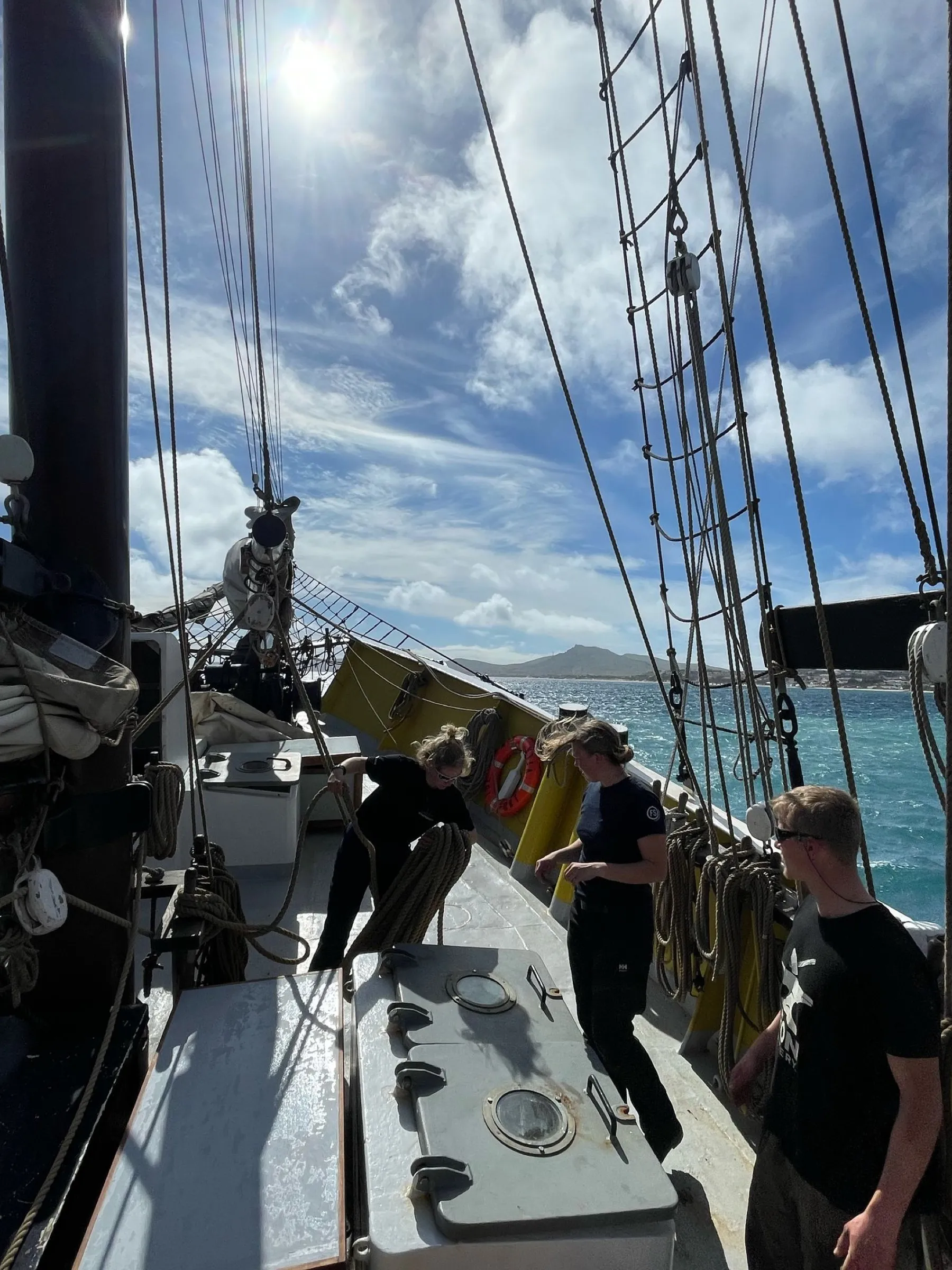 Drie bemanningsleden werken en schieten lijnen op aan dek in fel zonlicht terwijl het schip vertrekt, met de heuvels van Porto Santo en turkoois water naast zich