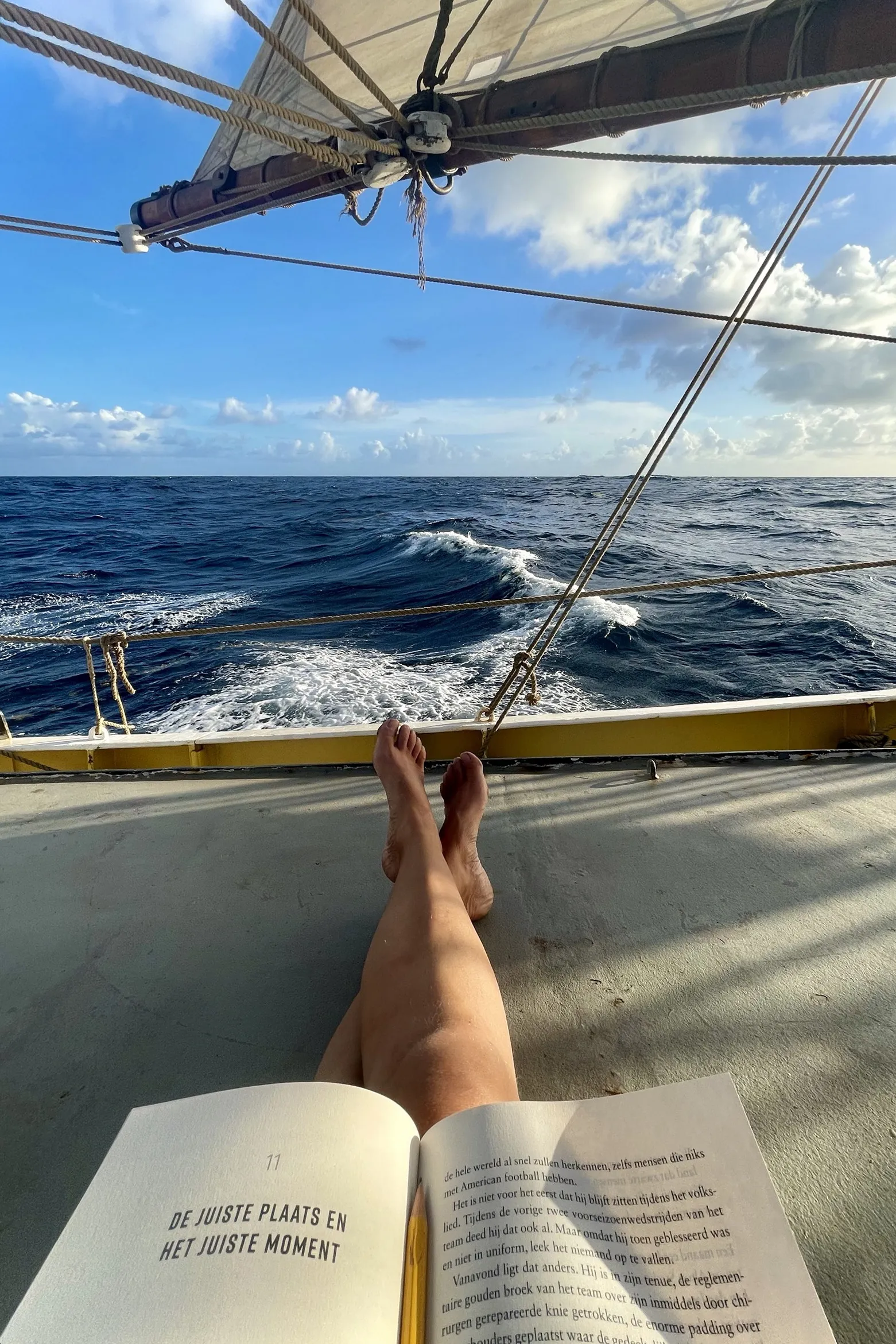 Bare legs stretched out on the warm deck with an open book resting on them, the sail overhead and the ship's wake running past the rail, blue water to the horizon