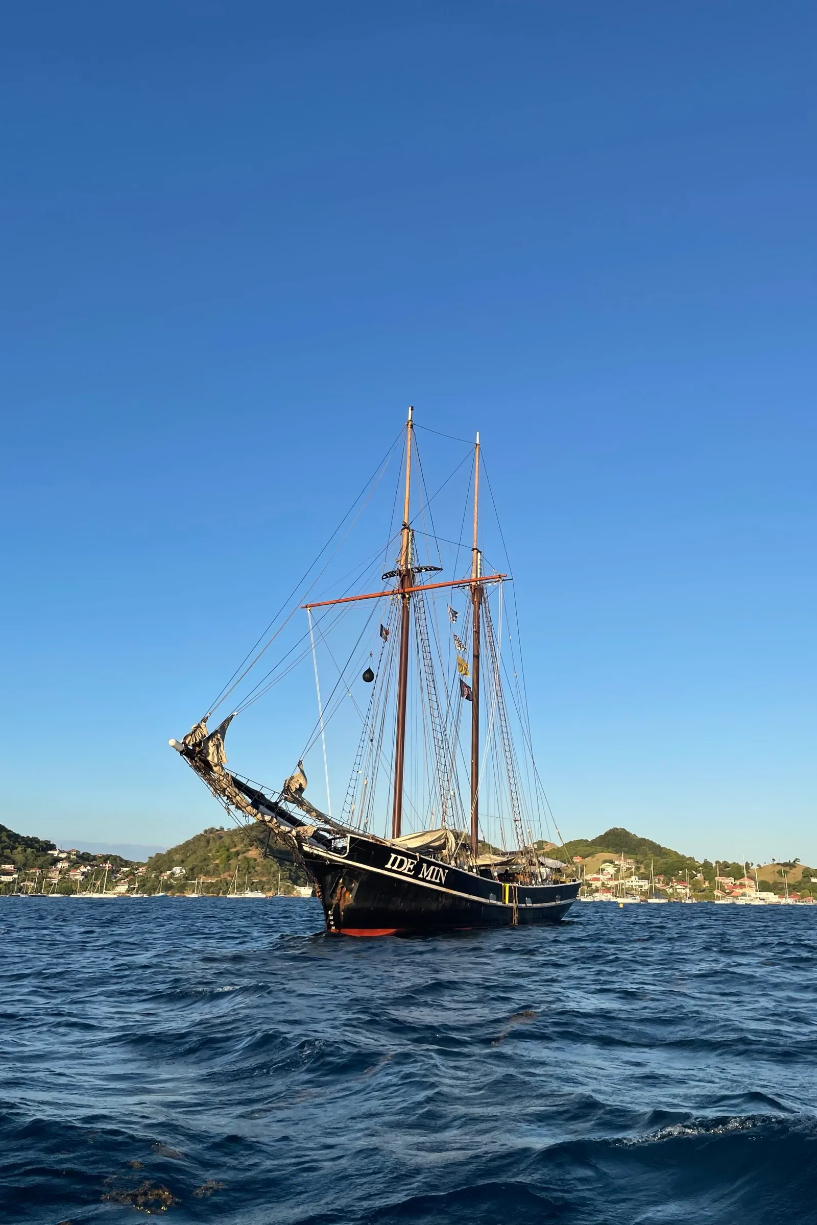 Ide Min voor anker in een Caribische baai, met haar naam op de boeg, groene heuvels en een kleine haven vol jachten achter zich